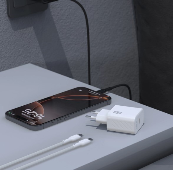 Ładowarka Dudao 30W GaN na szafce nocnej Biała ładowarka Dudao 30W GaN i kabel USB-C leżące na szafce nocnej obok ładującego się telefonu.