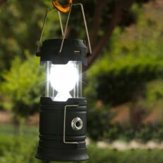 Rozsunięta lampa kempingowa Retoo wisząca na karabińczyku i świecąca dookoła, w tle zieleń