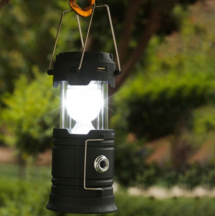 Rozsunięta lampa kempingowa Retoo wisząca na karabińczyku i świecąca dookoła, w tle zieleń